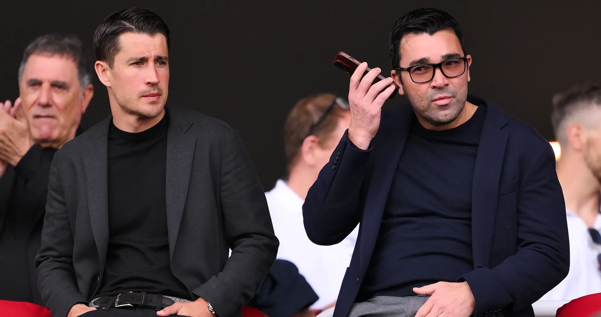 Two men seated in a sports venue, one on phone