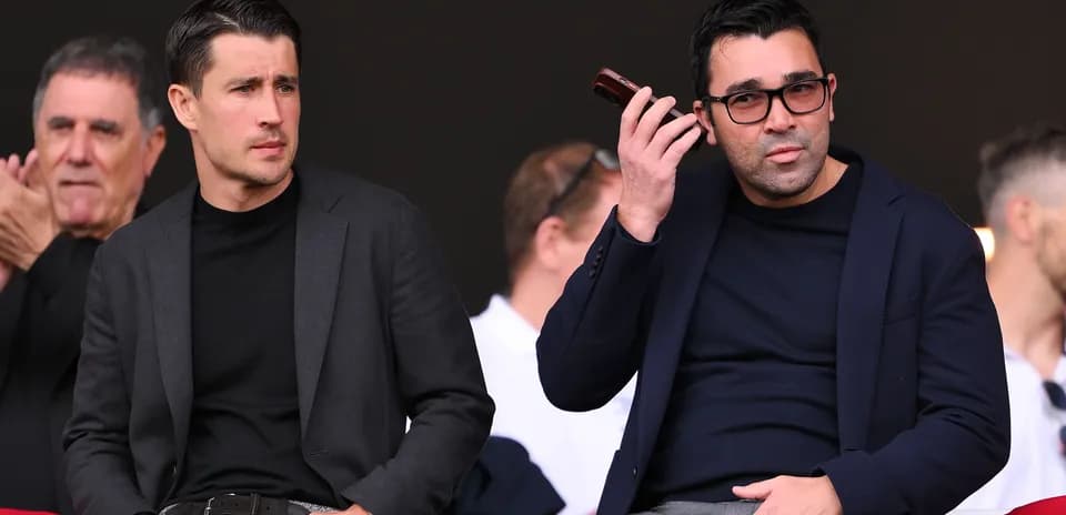 Two men seated in a sports venue, one on phone
